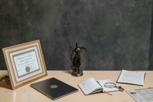 A lawyer's desk featuring a justice statue, diploma, laptop, magnifying glass, and documents.